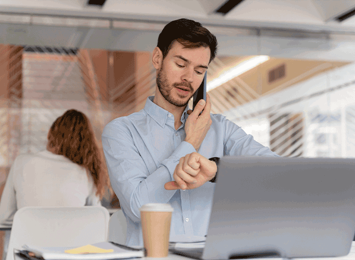 Um homem sentado à mesa falando ao celular enquanto olha para o relógio