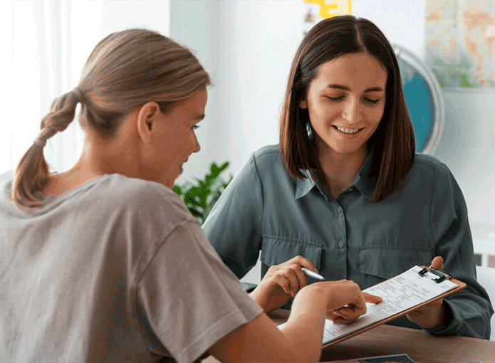 Duas mulheres sentadas e sorrindo, enquanto uma está com uma prancheta e uma caneta na mão, a outra aponta para um dado no papel